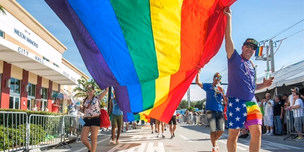A massive crowd of smiling people marching down a sunny street during a pride parade, collectively holding up a quarter-mile-long rainbow streamer that stretches into the distance.